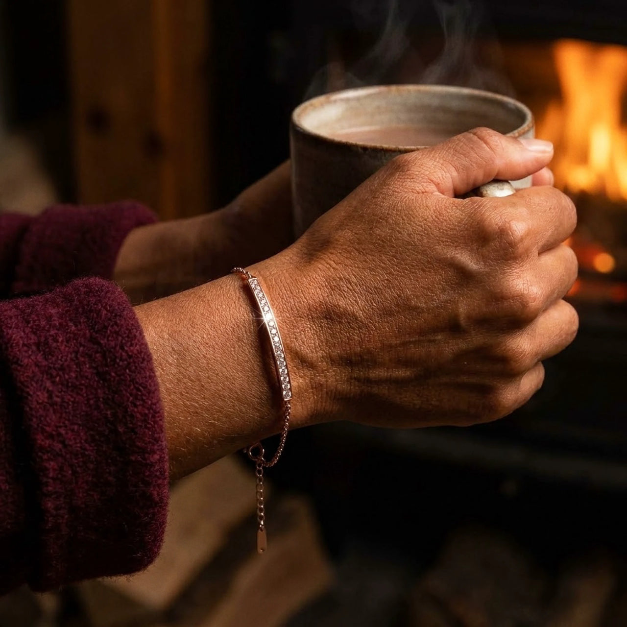 Crystal Bar Bracelet or rose porte au poignet, mains autour d'une tasse devant un feu
