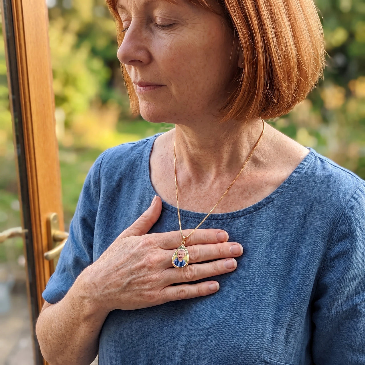 Woman in a blue linen shirt with her hand on her heart, wearing the gold secret drop photo necklace | SKU: TPH-DRPN-GOLD